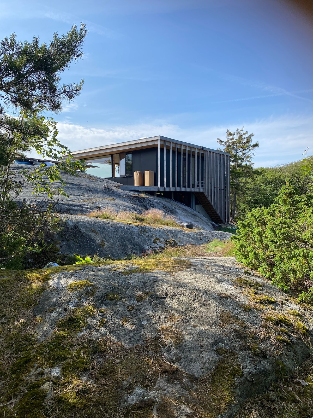 A modern cabin structure is nestled on rocky terrain, surrounded by lush greenery. The building features large windows and a covered deck, blending with the natural landscape. Sunlight brightens the exterior, highlighting the contrast between the wooden walls and the rocky surroundings.