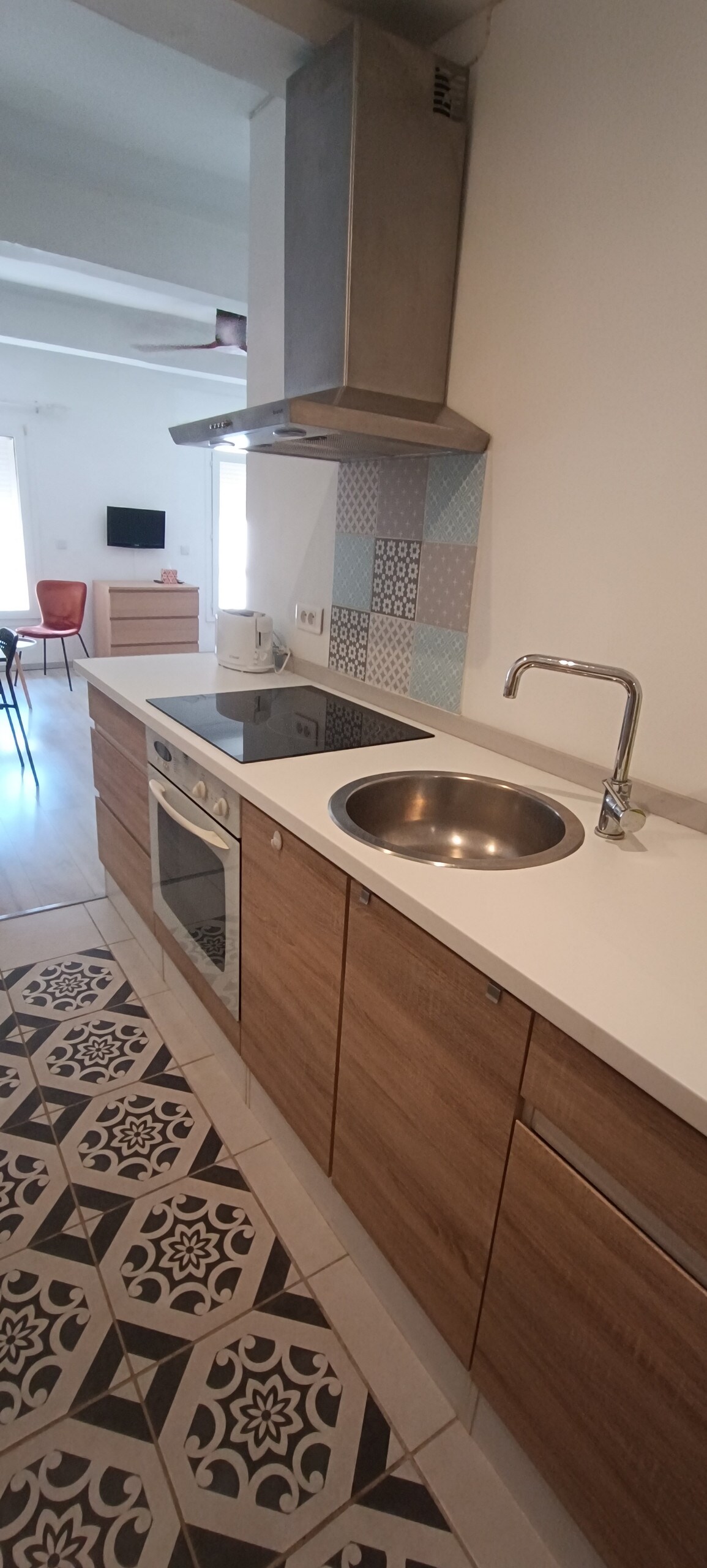 A kitchenette is shown featuring wooden cabinetry and a white countertop. A stainless steel sink and an electric stove are visible. The floor displays a patterned tile design, while light pours in from a nearby window, illuminating the space.