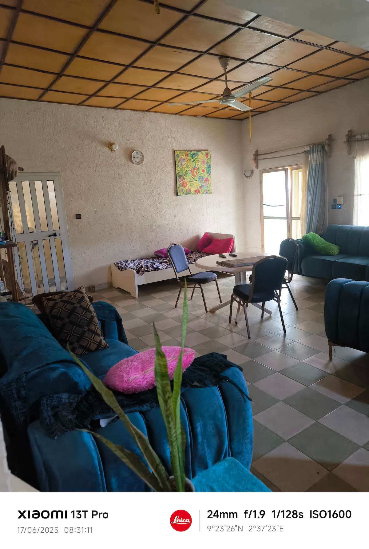 A living area is presented with soft blue seating and brightly colored cushions. Natural light enters through a window covered by sheer curtains. A small table stands on the floor, which features a tiled pattern. Art decorates the wall, providing a touch of color.
