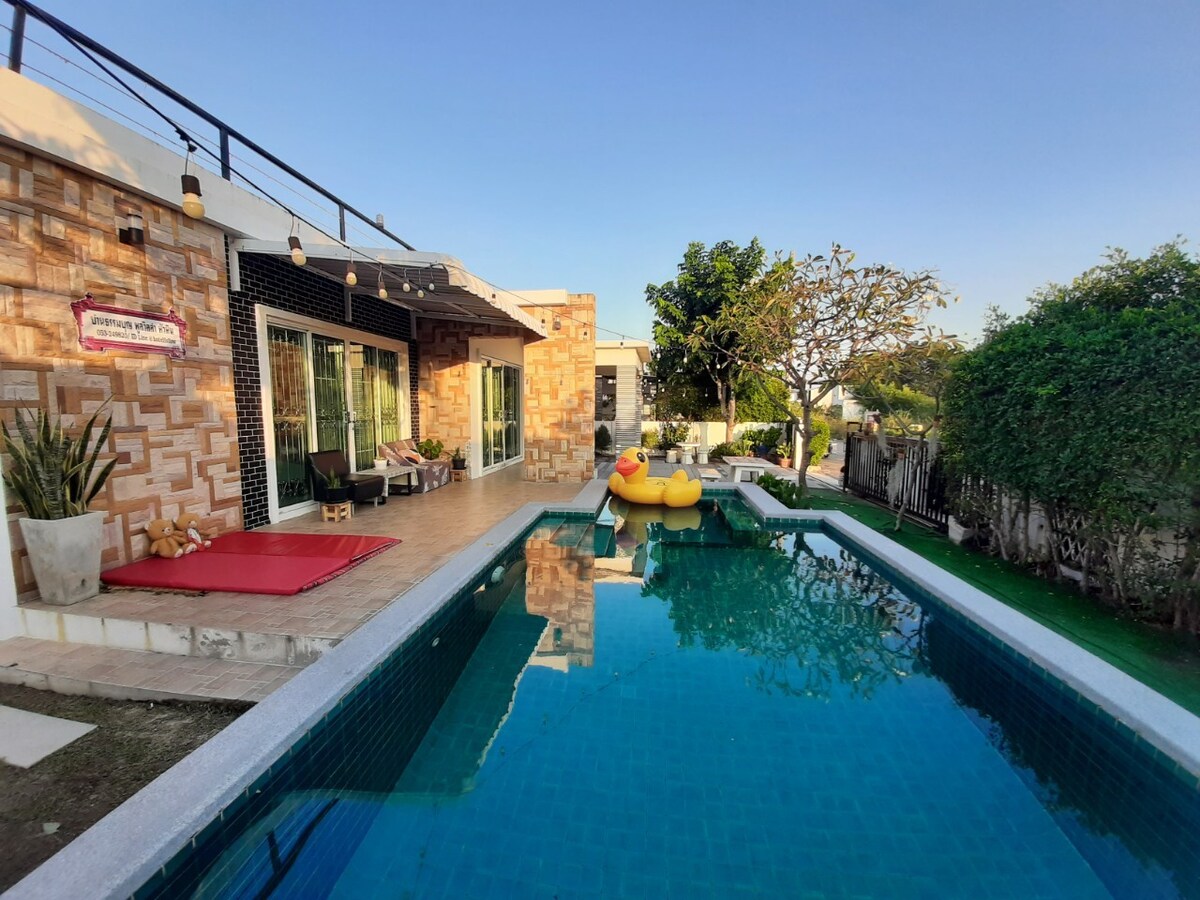 An outdoor area features a private swimming pool surrounded by green grass and a tiled patio. Lounge chairs are placed near the water, and a vibrant inflatable pool float adds a touch of color. The house is designed with large glass doors, allowing natural light to fill the space.
