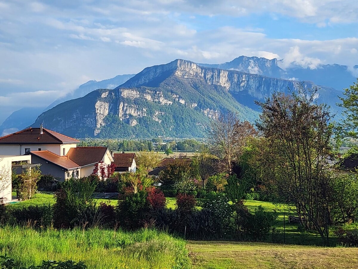 A view of rolling hills and a distant mountain range is showcased, highlighted by patches of green grass and trees. The landscape is dotted with houses featuring terracotta roofs, set against a backdrop of cloudy blue skies.