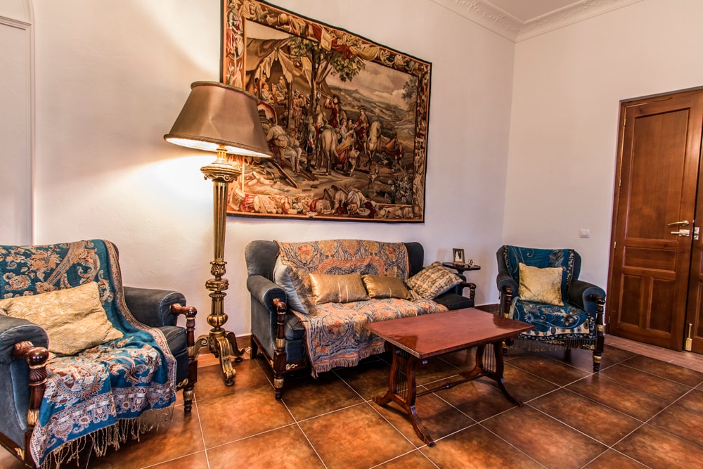 A common lounge area features two upholstered armchairs and a matching sofa, all adorned with decorative throws. A wooden coffee table sits in the center. A tall lamp provides soft illumination, while a detailed tapestry hangs on the wall, enhancing the room's character.