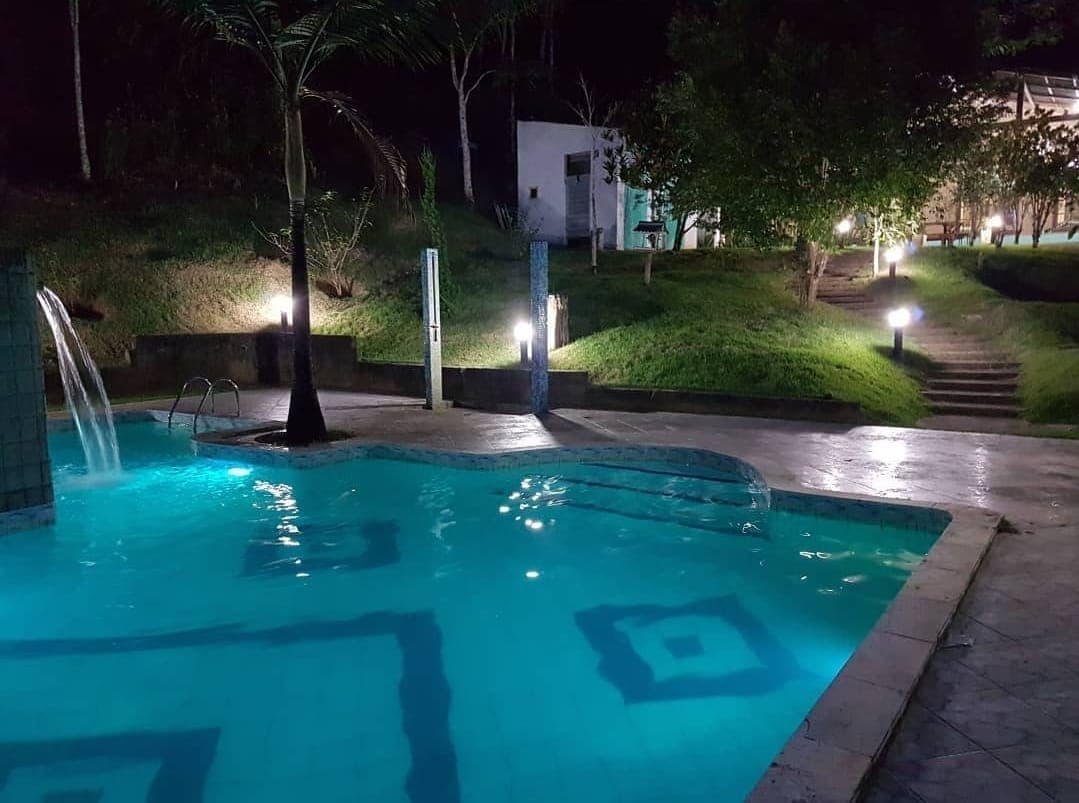 An illuminated outdoor area features a swimming pool with a gentle waterfall, surrounded by greenery. Pathways lead to a nearby building, while soft lighting enhances the tranquil ambiance. The design of the pool includes geometric patterns that stand out against the clear water.