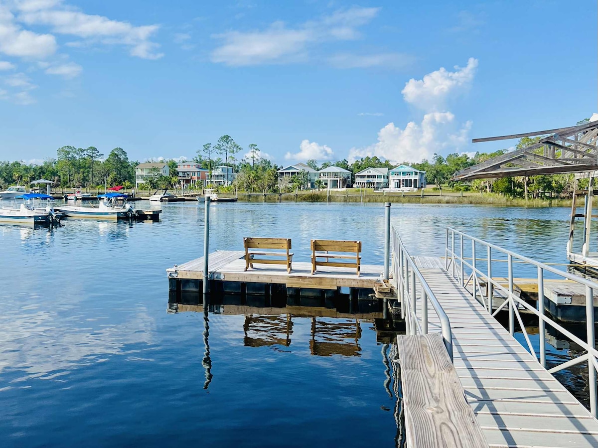 A serene dock area is depicted, featuring wooden benches positioned to overlook the water. Small boats are anchored in the background, with lush greenery lining the shore. Clear blue skies reflect on the calm surface, creating a tranquil environment.