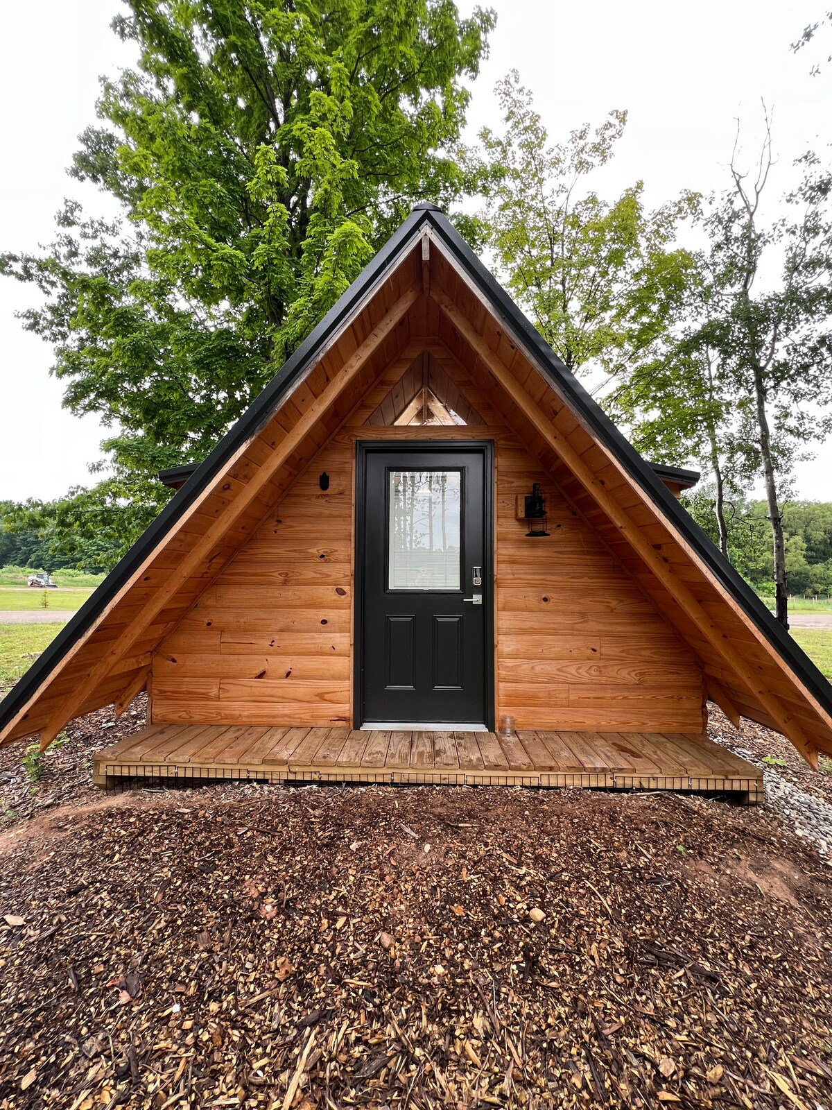 The front exterior of a rustic A-frame cabin is shown, constructed from wood with a natural finish. A dark front door is centered under a small overhang. The entrance is flanked by a lantern and surrounded by a wooden deck, with mulch and greenery in the foreground.