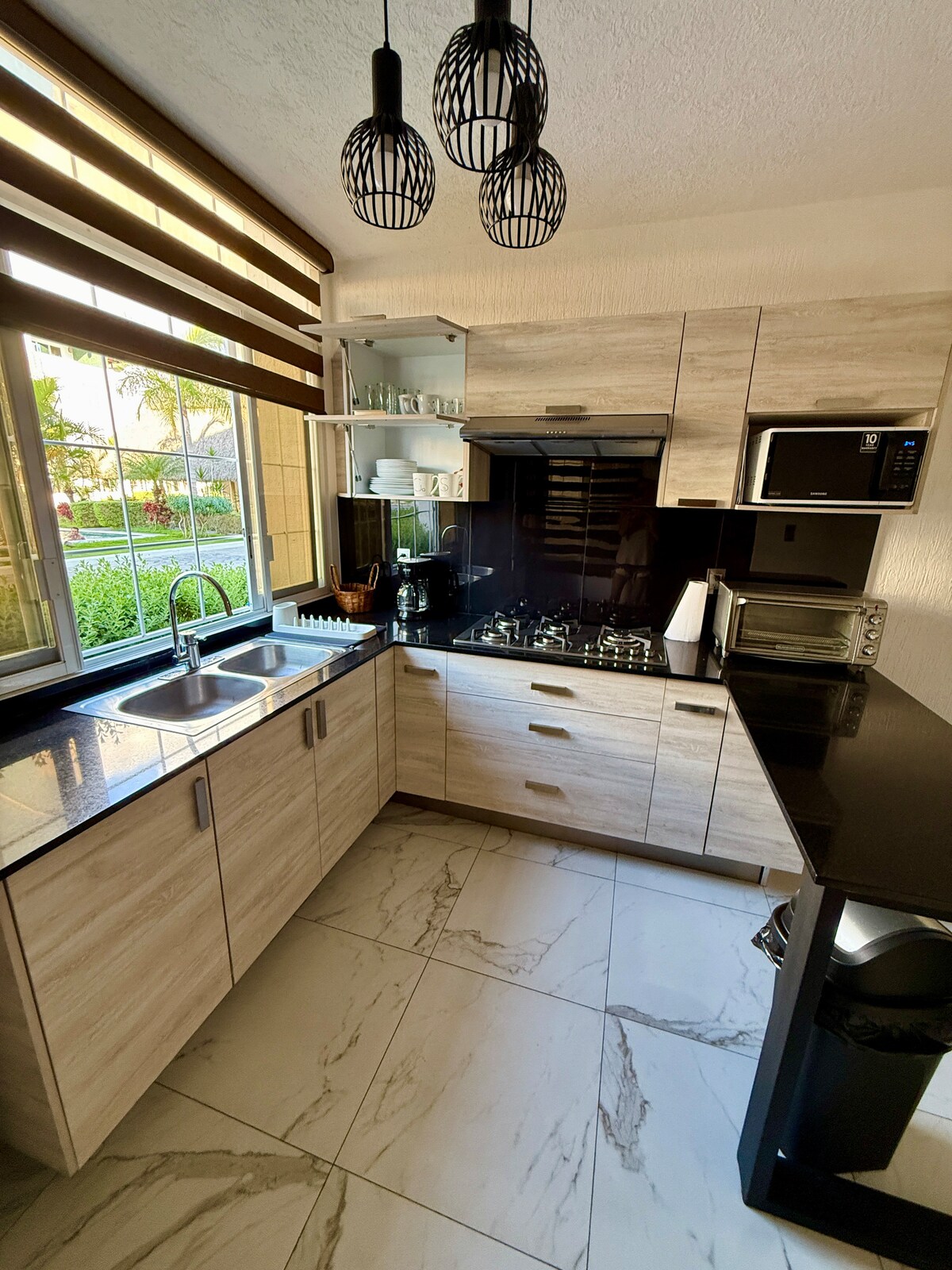 A modern kitchen is depicted, featuring sleek cabinets and a black countertop. A stainless steel sink is positioned under a large window that offers a view of the greenery outside. Kitchen appliances, including a stovetop and microwave, are neatly arranged for functional use.