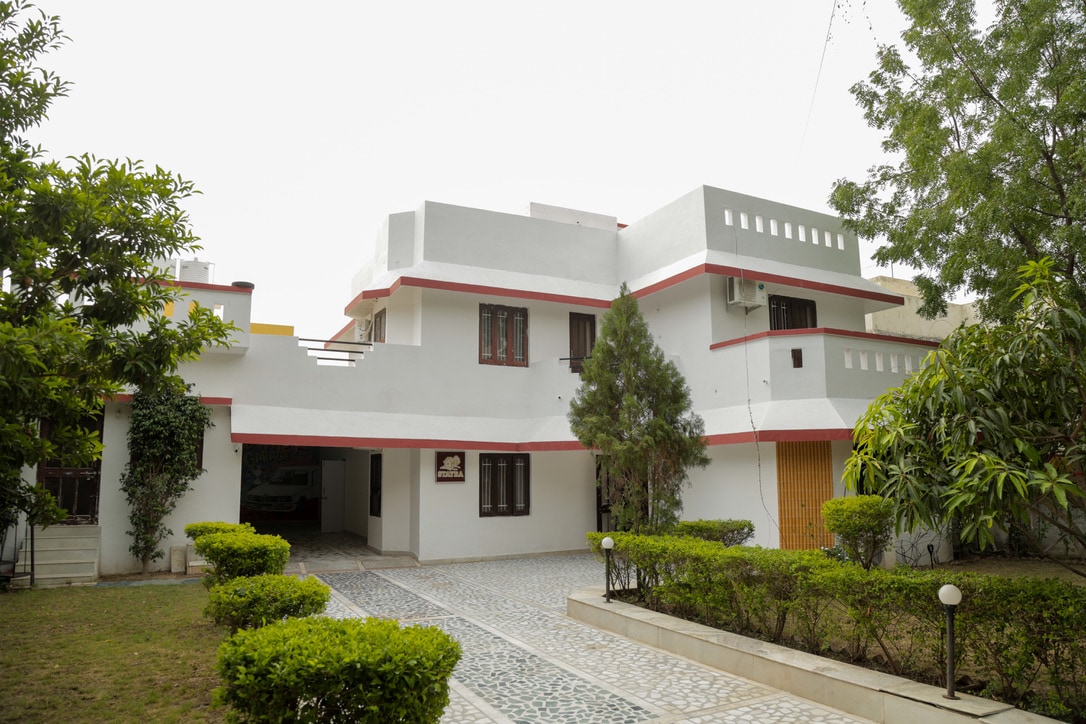 The villa's exterior is highlighted with a clean, modern design featuring a two-story structure. Lush greenery surrounds the property, with well-maintained shrubs and a cobblestone pathway leading to the entrance. Large windows allow ample natural light, enhancing the inviting ambiance.