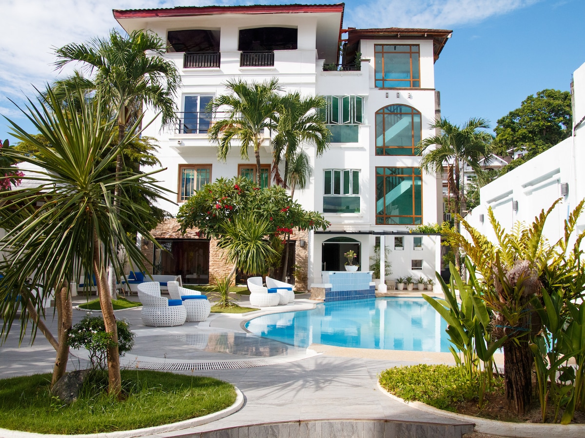 A bright and inviting view of the property’s exterior, featuring a large swimming pool surrounded by lush tropical landscaping. Comfortable seating areas are situated poolside and the three-story building displays numerous windows, allowing ample natural light to fill the space.