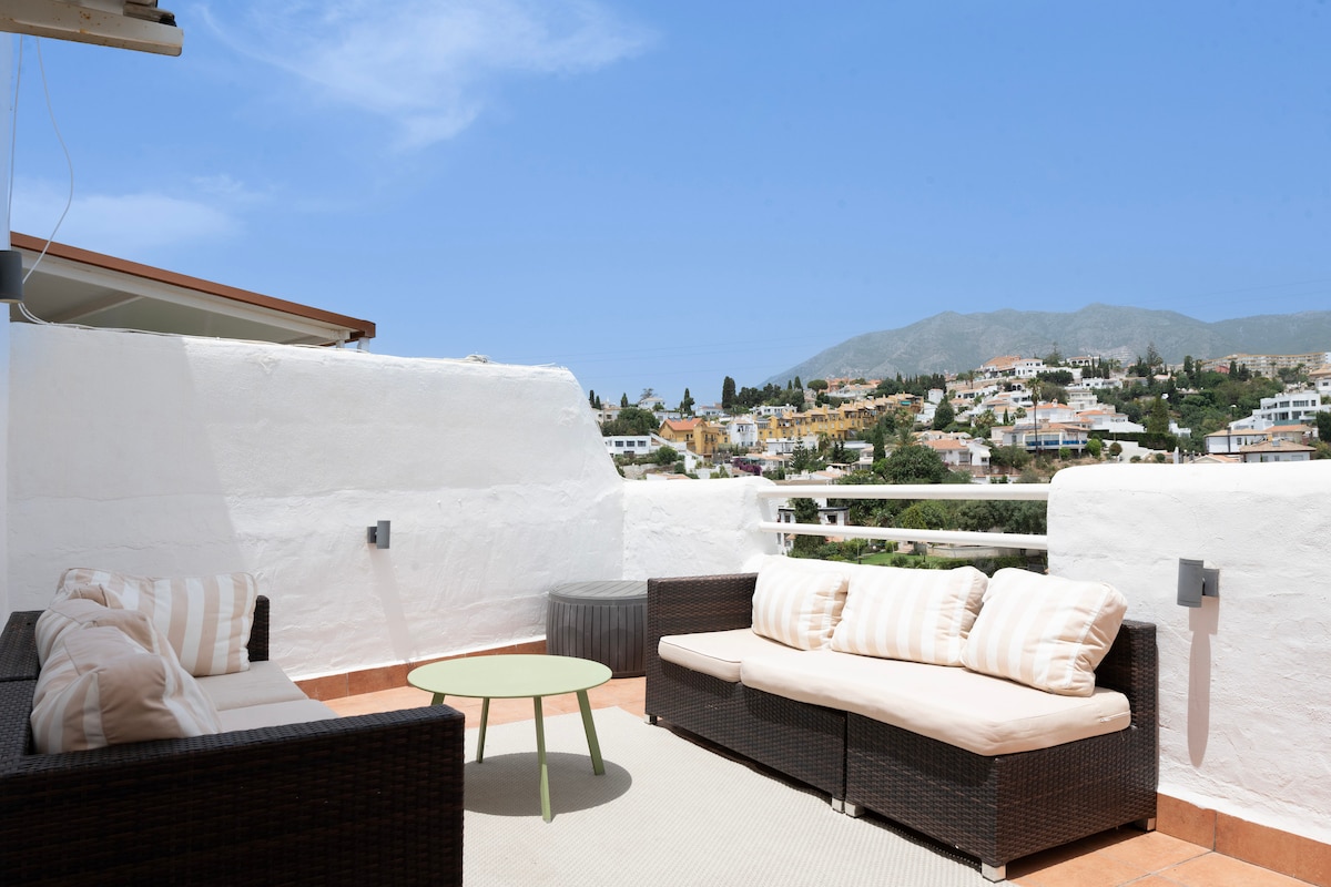 A private terrace features a seating arrangement with a wicker sofa and light cushions, complemented by a small round table. The surrounding view includes distant mountains and a residential area, all under a clear blue sky.