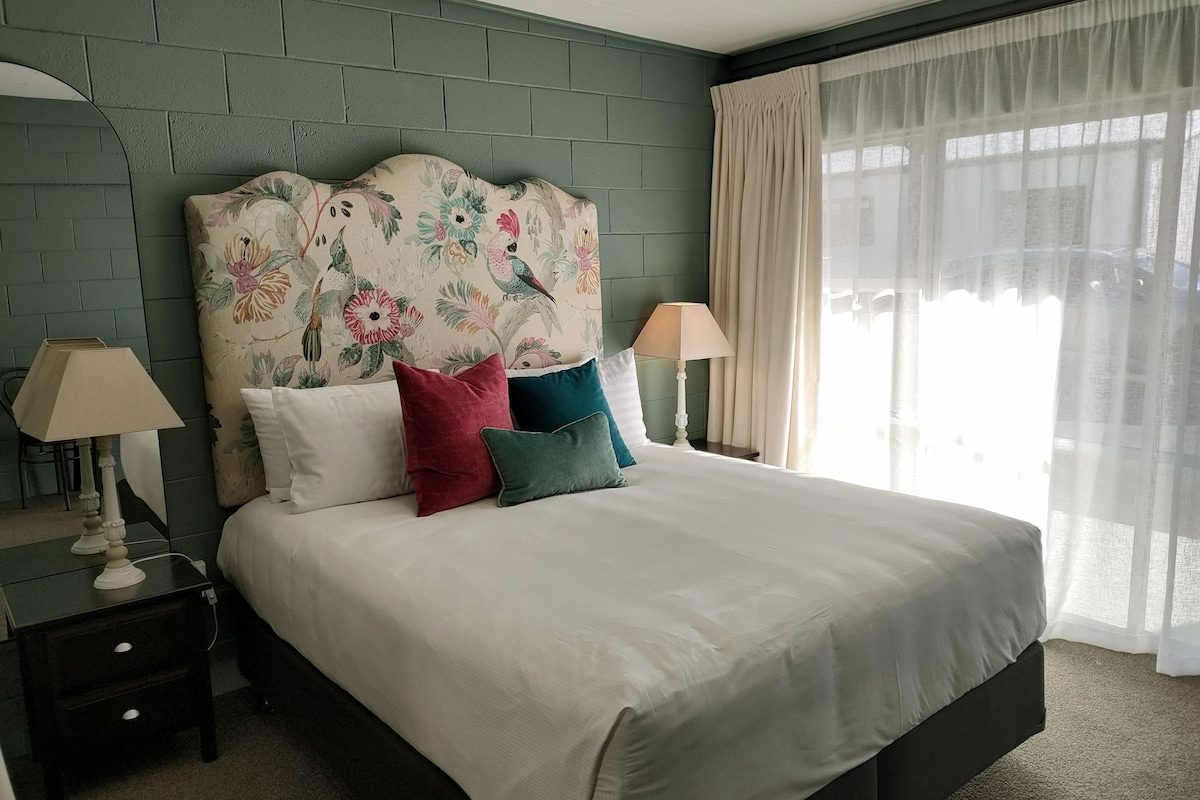 A cozy sleeping area is highlighted by an upholstered headboard featuring floral and bird designs. Neutral bedding is complemented by decorative cushions in shades of green and burgundy. Two bedside lamps sit on dark wood nightstands, and natural light filters through sheer curtains.