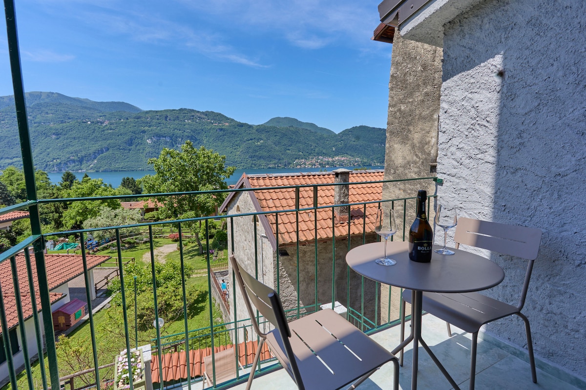 A small balcony is depicted, featuring a round table with two chairs. Views of the surrounding mountainous landscape and the lake are visible, with sunlight illuminating the area. The balcony's railing adds a welcoming touch to the outdoor space.