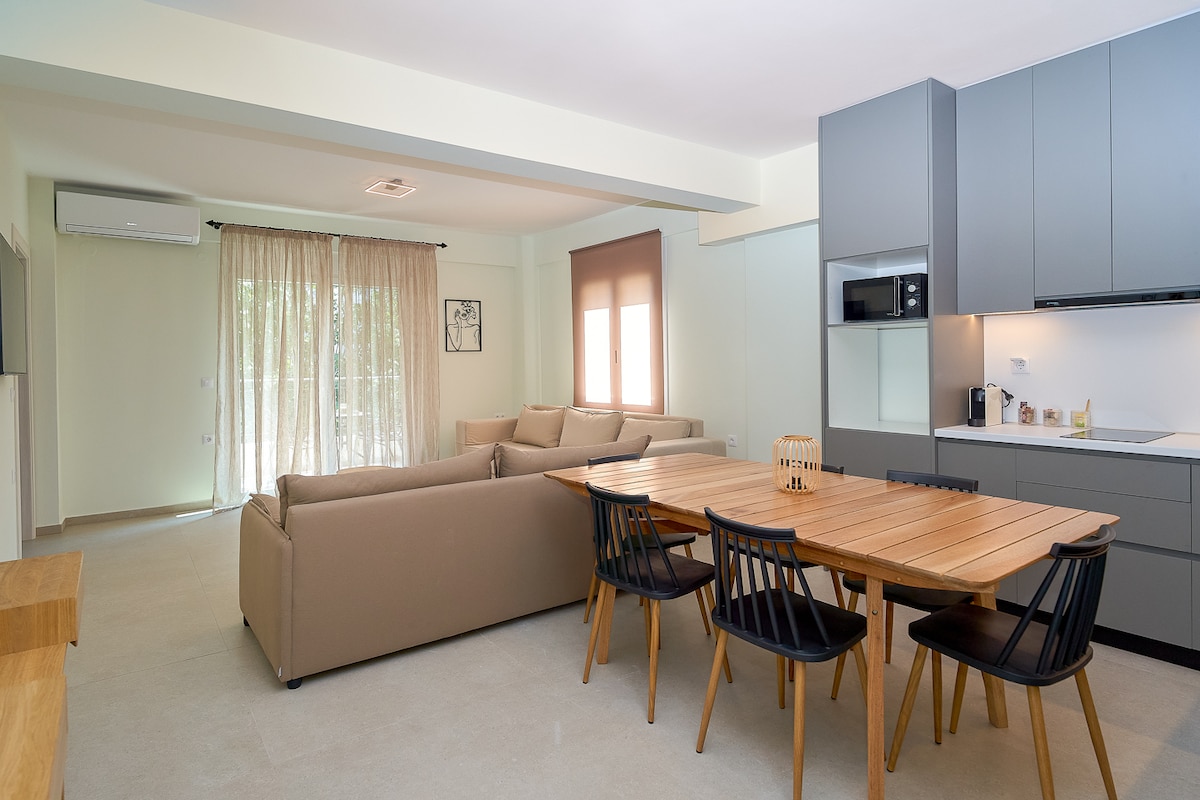 An open-plan living and dining area features a light wood dining table surrounded by black chairs. A spacious beige sofa is visible, positioned near large windows with sheer curtains, allowing for natural light to brighten the space. Modern kitchen appliances are seen in the background.