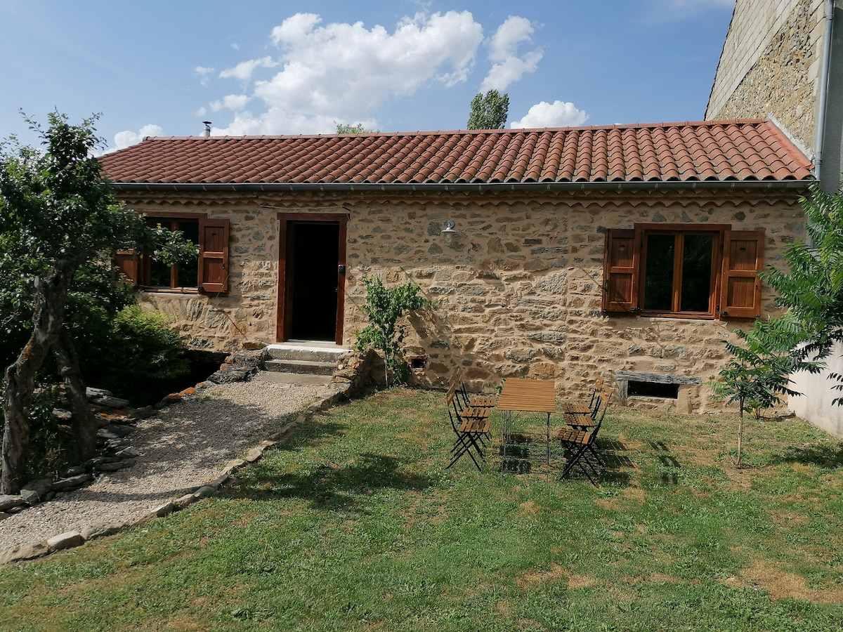 A rustic stone cottage is surrounded by a green lawn, accented by trees and shrubs. Two metal chairs and a small table are situated on a gravel pathway, leading to a wooden door that opens onto a welcoming entrance. Natural light filters through the windows.