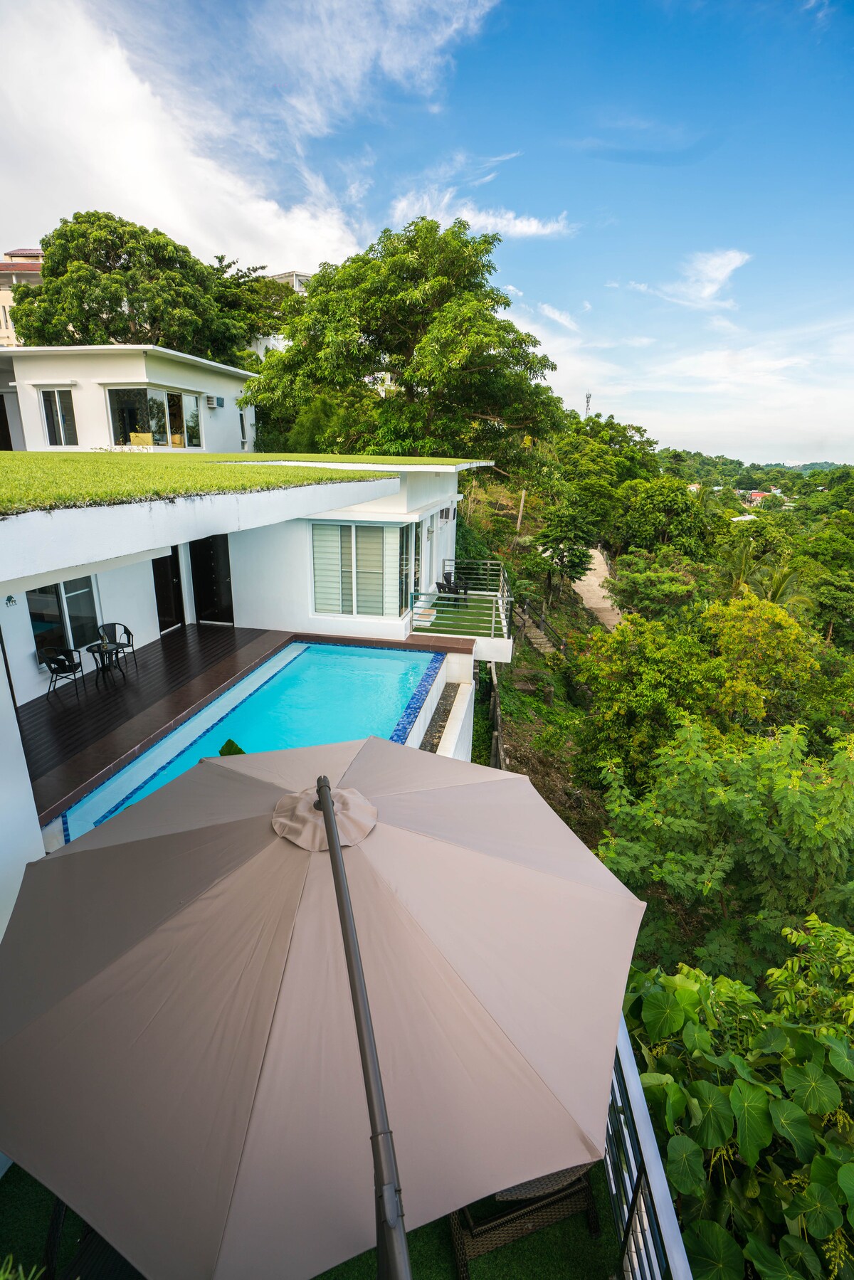 A spacious villa is positioned on a hillside, featuring a private pool bordered by a wooden deck. An umbrella provides shade over seating arrangements, and lush greenery surrounds the property, enhancing the natural setting. A clear blue sky is visible above the villa.