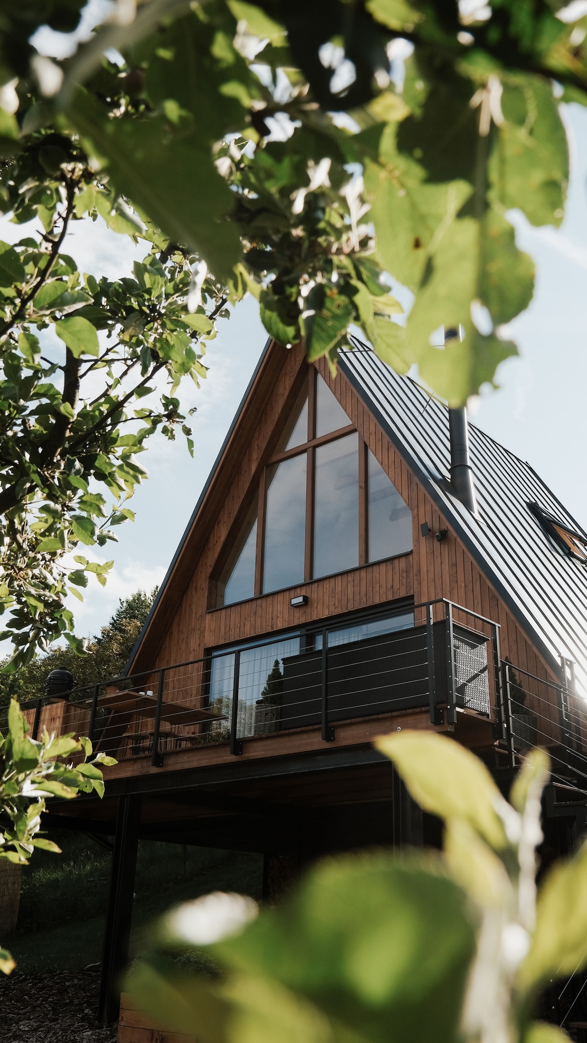A modern wooden structure features an A-frame design with large windows that allow natural light to flood the interior. The deck is surrounded by greenery, providing a peaceful setting amidst nature. The architecture emphasizes a clean, contemporary aesthetic.