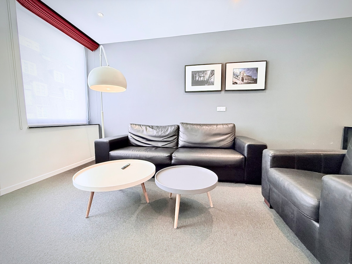A cozy living area is featured, complete with a dark leather sofa and an armchair. Two coffee tables in contrasting colors are placed in front, and framed black-and-white photographs adorn the wall. Natural light filters through the partially covered window.