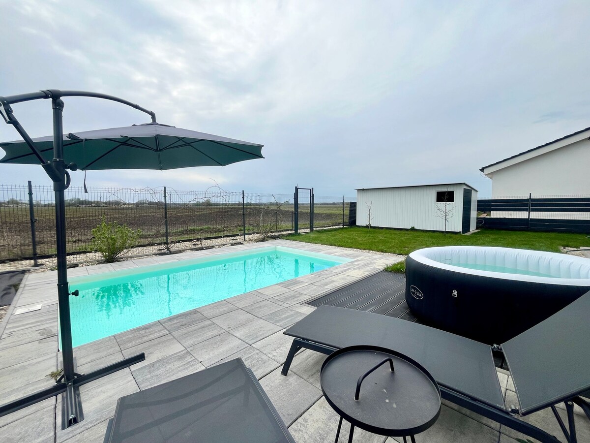 A tranquil outdoor area features a clear swimming pool surrounded by a paved patio. Comfortable lounge chairs with a small table sit nearby, and a shaded umbrella provides cover. A hot tub is positioned to one side, with a grassy area and a distant fenced property visible in the background.