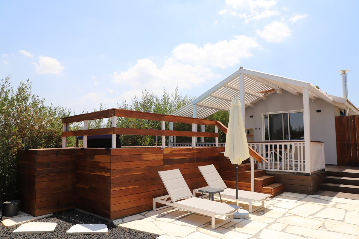 A spacious deck is featured with wooden fencing and lounge chairs under an umbrella. The structure is designed with large windows, offering natural light, while greenery surrounds the area, creating a serene outdoor environment.