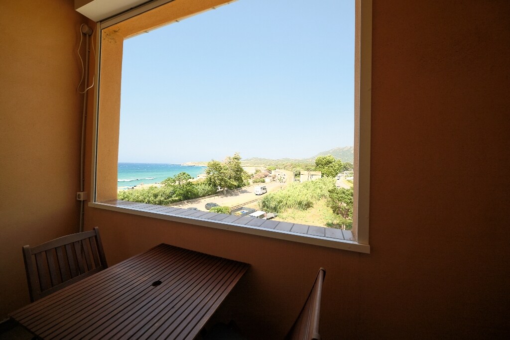 A large window offers a clear view of the beach and sea, framed by the room's warm walls. A wooden table with matching chairs is positioned in front of the window, inviting relaxation while enjoying the scenic coastal landscape outside.