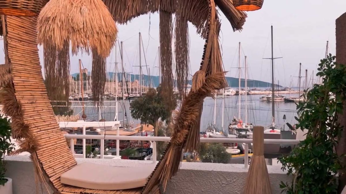 A unique woven swing chair is positioned on a balcony overlooking a marina filled with yachts and sailboats. The soft, natural textures of the chair complement the serene waters and distant hills, creating a peaceful space for relaxation.