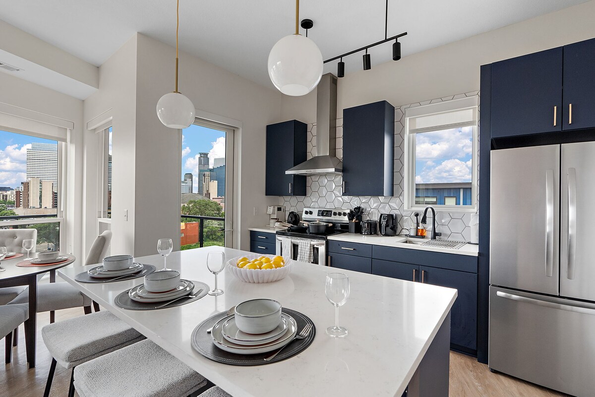 A modern kitchen is presented, featuring stylish navy cabinetry and a large white island with seating. Stainless steel appliances, including a refrigerator and oven, are visible. Large windows frame city views, while the dining table is set with plates and glasses, inviting shared meals.