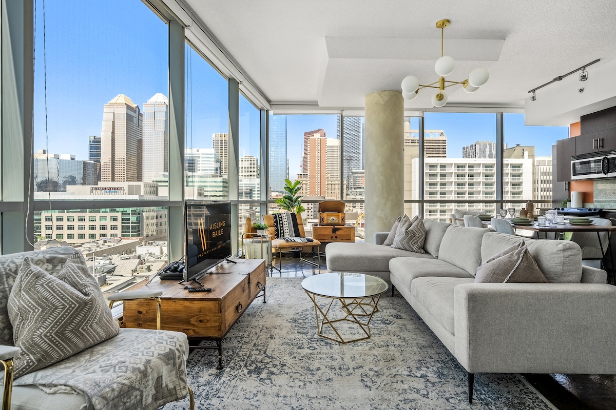 A spacious living area features floor-to-ceiling windows that offer views of the downtown Calgary skyline. A light-colored sectional sofa is paired with a glass coffee table and wooden accents. A 55" Smart TV is positioned on a wooden console, enhancing the room's modern decor.