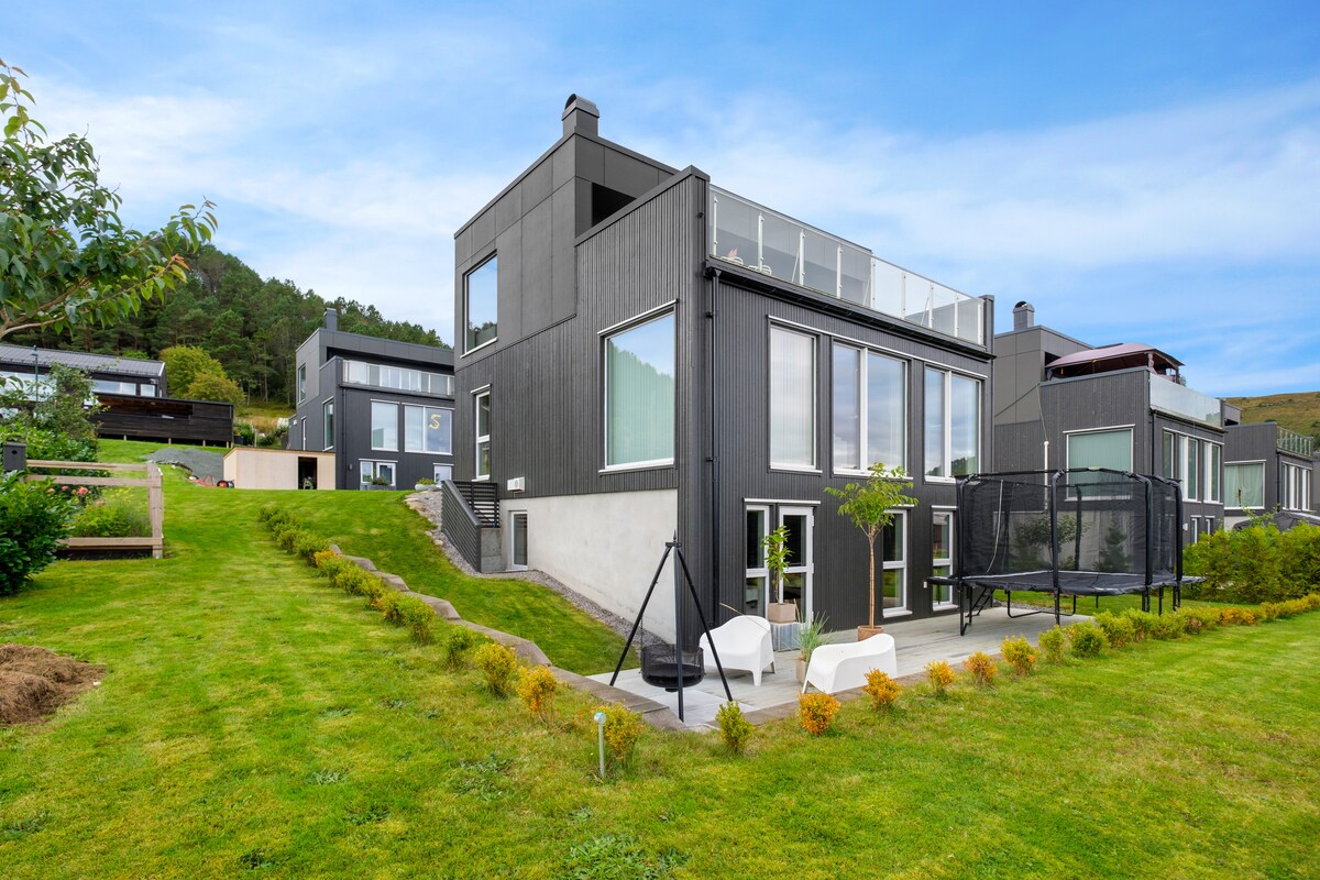 A modern detached house is showcased, featuring sleek black exterior walls and large windows that allow ample natural light. A spacious yard is visible, complete with landscaped greenery and patio furniture. The surrounding area includes other contemporary homes, highlighting a peaceful and family-friendly environment.