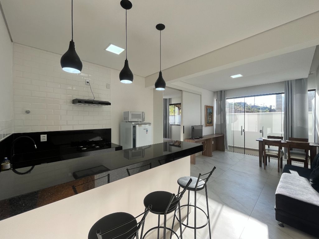 An open-plan kitchen and living area is shown, featuring a polished black countertop and modern appliances. Three pendant lights hang above a seating area with three stools. A wooden dining table and chairs are positioned nearby, while large windows allow natural light to flood the space.