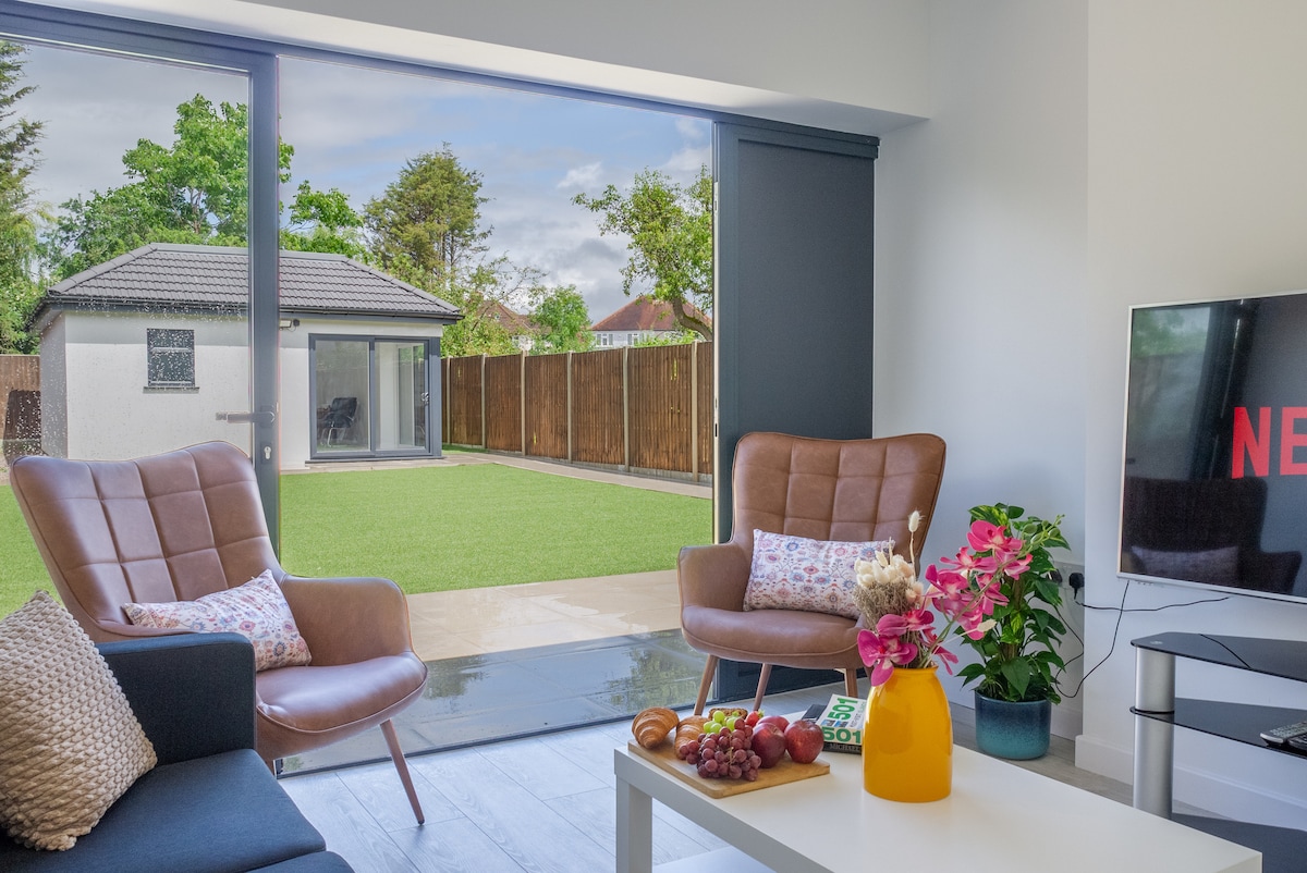 A bright living area showcases two comfortable armchairs, facing large glass doors that open to a private garden. The space includes a sleek table with fruit, pastries, and a television with the Netflix logo. Light enters through the doors, enhancing the airy atmosphere.