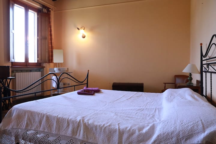 غرفة كبيرة بإطلالة رائعة (نوافذ معتمة) مجهزة بخزانة، وصندوق، وكرسي مع طاولة، وطاولات جانبية مع باب ودرج وسرير مزدوج. سقف مرتفع جدًا مع عوارض وطوب مكشوف.