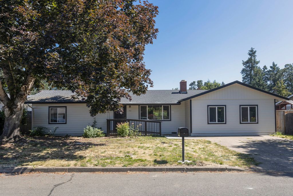 A single-story home features a wide front porch with steps leading to the entrance. Lush greenery surrounds the property, complemented by a large tree providing shade. A driveway and curbside parking area are visible, along with multiple windows allowing natural light into the interior.