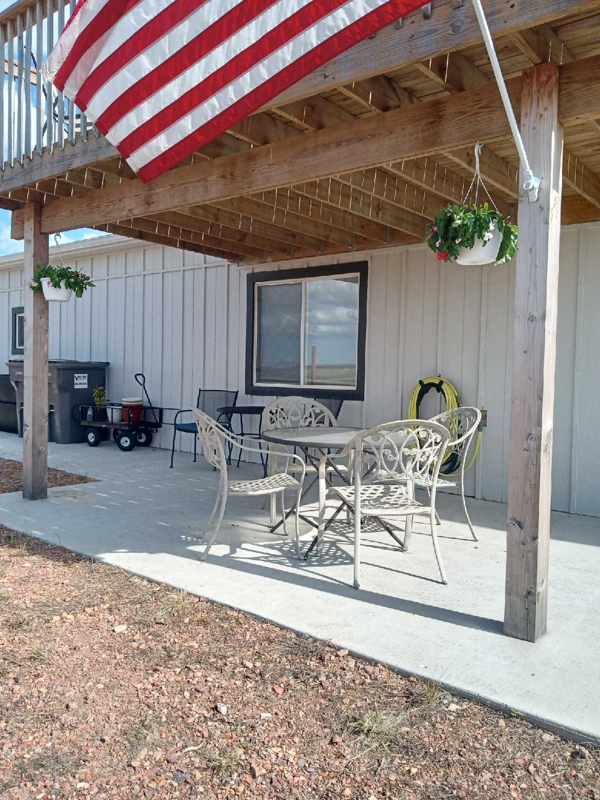 An outdoor patio area is shown, featuring a metal table surrounded by four chairs. Two hanging planters are positioned above the table, and a flag is displayed nearby. A utility cart and garden hose are visible, with a large window providing natural light.