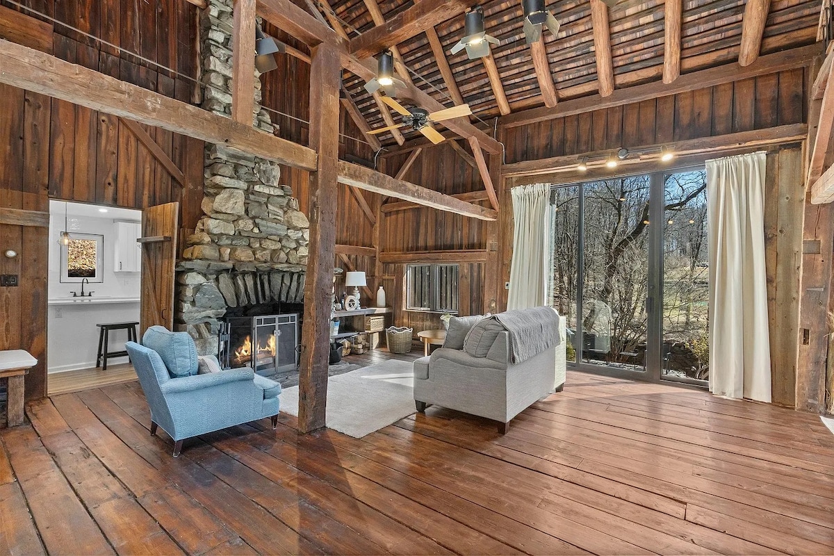 A spacious living area featuring a stone fireplace with a wooden mantel. Large windows allow natural light to fill the room, highlighting the wooden beams and floor. A comfortable couch and a blue armchair create a cozy seating arrangement, surrounded by a minimalist design.
