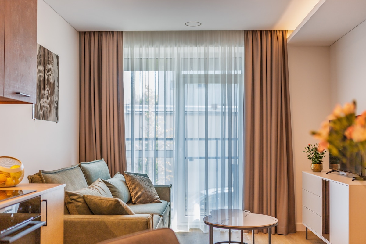 A light-filled living area is showcased with large windows dressed in sheer curtains, allowing for natural illumination. A comfortable sofa with several cushions offers a cozy seating option. A small coffee table is positioned in front of the sofa, complemented by a console with decorative items.