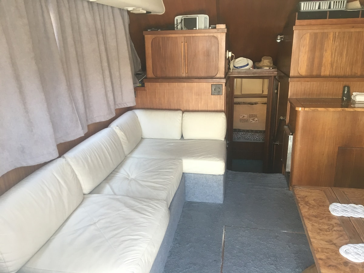 A light-colored sectional sofa is positioned in a cozy seating area. Dark wood cabinetry lines the walls, and soft, gray curtains frame the window, diffusing natural light. A wooden table is seen to the right, enhancing the space's functionality.
