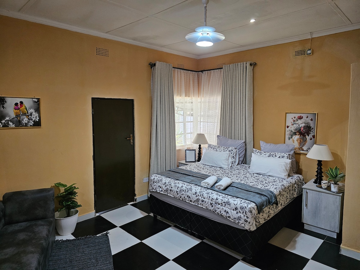 A king-size bed is centrally placed, adorned with crisp linen and two rolled towels. A small side table with a lamp is positioned beside the bed. Natural light filters through sheer curtains, illuminating the room, which features a cozy seating area with a grey sofa and decorative plants.