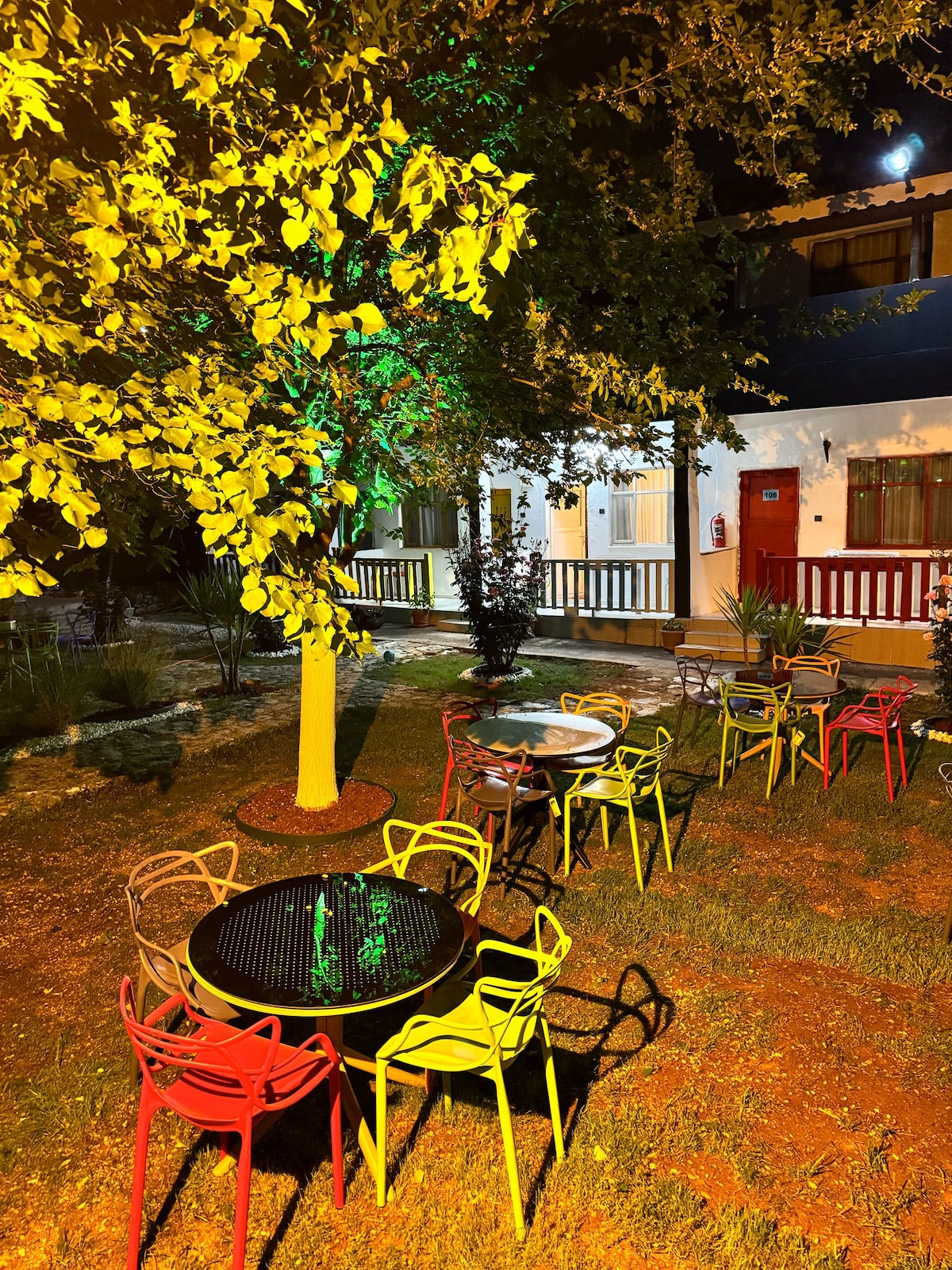 An outdoor area is presented with several colorful chairs surrounding small tables on a grassy surface. Soft lighting highlights the foliage of nearby trees, creating a relaxed ambiance. The building in the background features a white façade with red accents and a welcoming atmosphere.