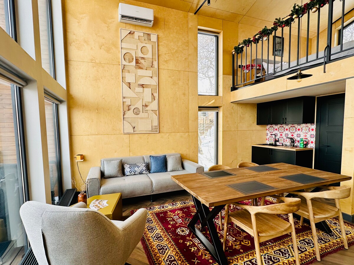 A spacious living area features large windows, allowing natural light to illuminate the interior. A comfortable gray sofa is accompanied by a stylish armchair and a wooden dining table with four chairs. Wall art adds character, and the kitchen area is visible in the background.