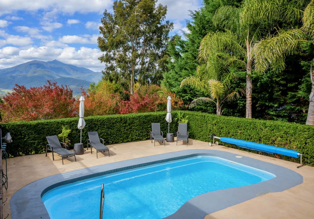 An outdoor saltwater pool is framed by lush greenery and colorful foliage. Sun loungers are positioned around the pool, ensuring a comfortable space for relaxation. Umbrellas provide shade, while a scenic mountain view is visible in the background beneath a partly cloudy sky.