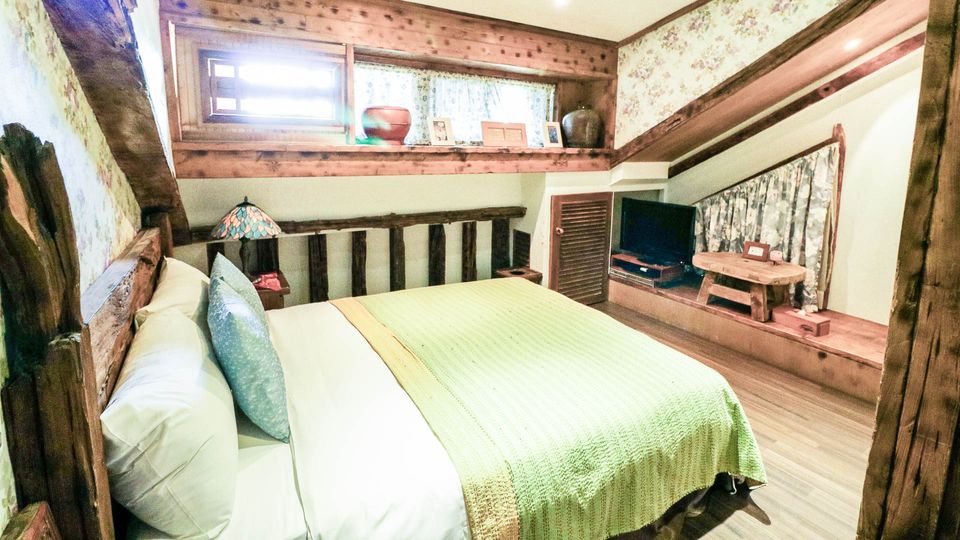 A cozy bedroom features a spacious bed covered with a light green blanket. Wooden accents line the walls, complemented by floral wallpaper. Natural light enters through the window, illuminating the area, while a television and small wooden table complete the inviting setting.