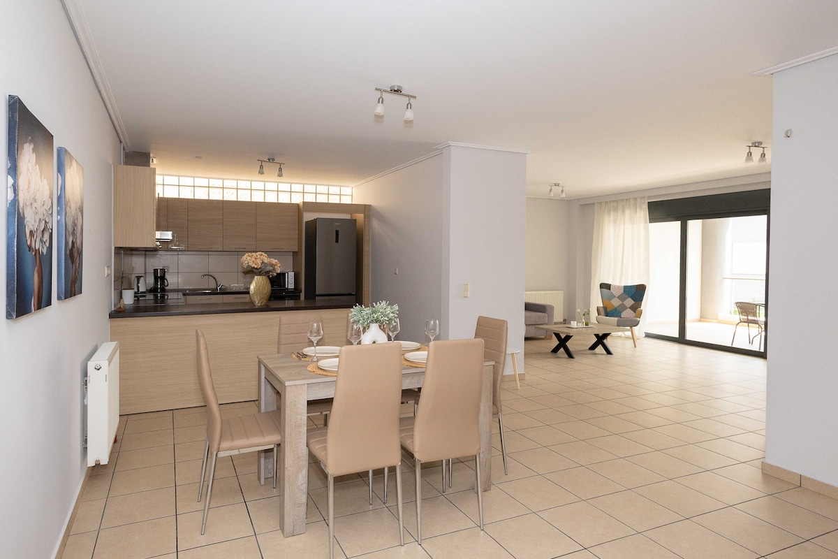 A spacious living area is depicted, featuring a modern dining table set for four. The kitchen is seamlessly integrated, showcasing wooden cabinetry and essential appliances. Natural light fills the room through large glass doors leading to an outdoor space, creating a bright and inviting environment.