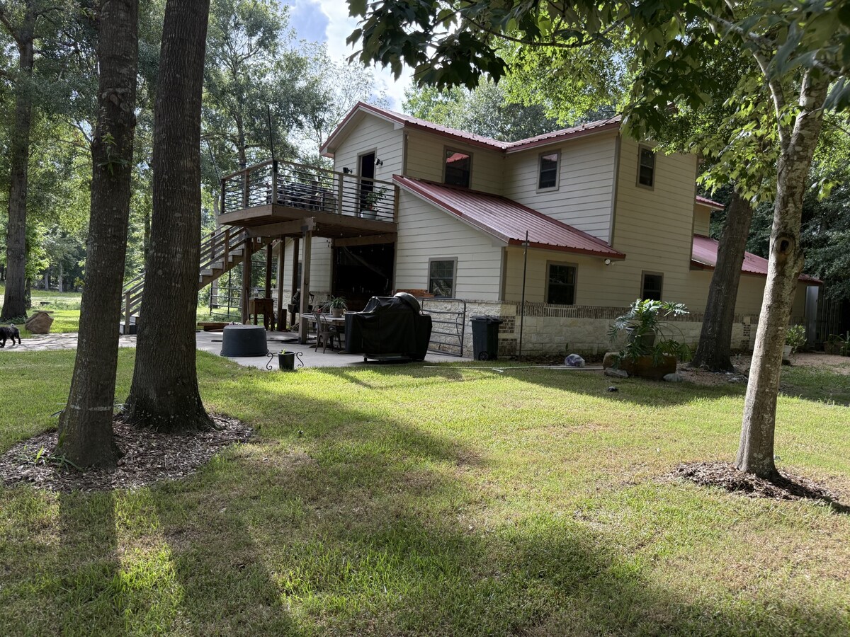 A two-story house is depicted, featuring a red metal roof and a spacious balcony above the patio. The surrounding area is landscaped with grass and mature trees. A fire pit and outdoor seating can be seen on the patio, indicating potential for outdoor gatherings.