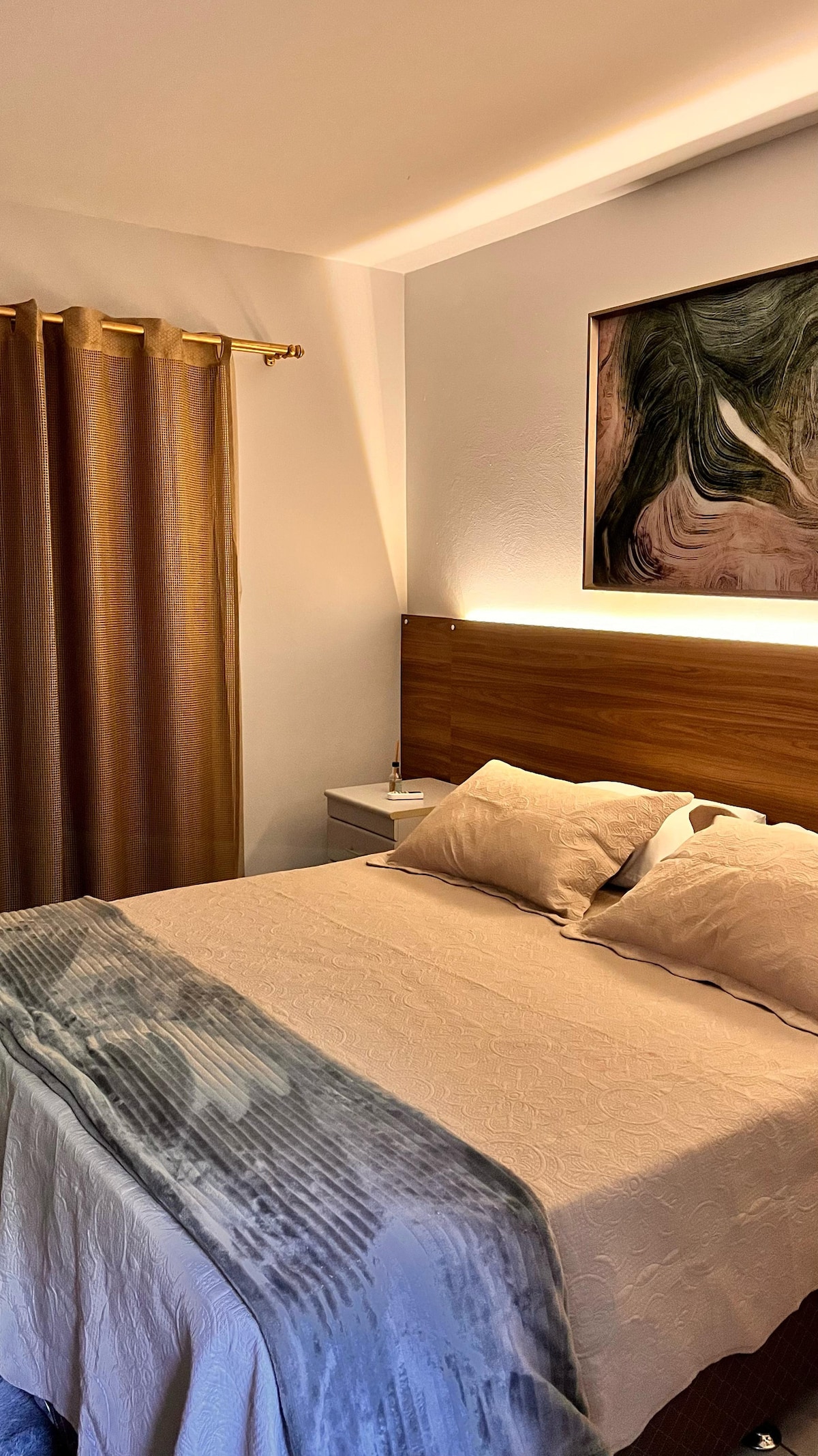 A cozy bedroom features a wooden headboard with integrated lighting above a spacious bed dressed in neutral linens. Two plump pillows rest at the head, while a soft throw is draped over the foot of the bed. A warm curtain complements the inviting atmosphere.