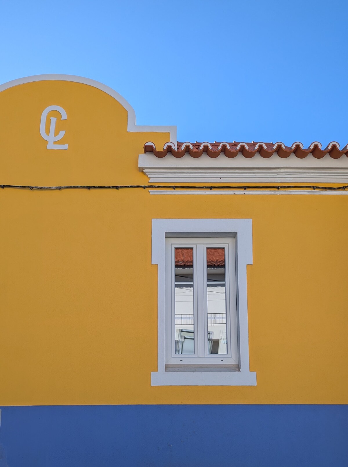 A bright yellow building features a white-framed window with three vertical panes. The roof is lined with red tiles, while the lower section contrasts in blue. Simple decorative elements include a curved top and a stylized design above the window, set against a clear blue sky.