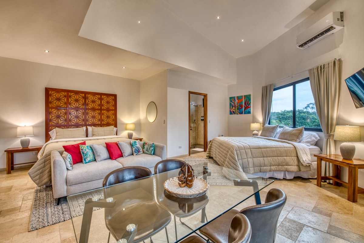A spacious room is presented with two king beds, each adorned with soft bedding. A dining area featuring a glass table and chairs offers a comfortable space for meals. Natural light fills the room through a large window, complemented by tasteful art on the walls.