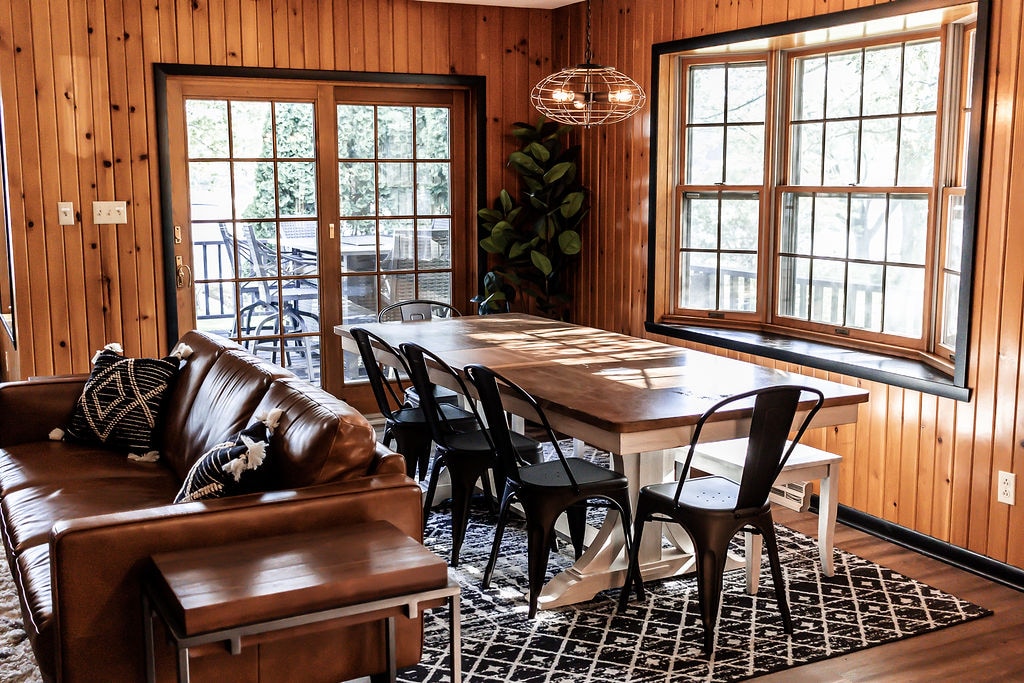 A spacious dining area features a large wooden table surrounded by black chairs. Light from multiple windows illuminates the room, which is accented by natural wood paneling and a cozy seating arrangement with a brown leather sofa. A stylish light fixture hangs above the table.