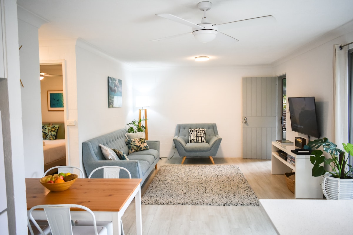 A spacious living area features modern furnishings, including a comfortable blue sofa and an accent chair. Natural light fills the room, highlighting a vibrant area rug. A small dining table with fruit sits nearby, and a television on a console adds to the cozy atmosphere.