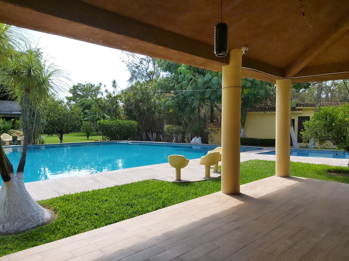 A large outdoor pool is framed by lush greenery and neatly trimmed grass. Patio seating made of yellow materials is positioned under a sturdy overhang. The serene environment is complemented by distant trees, creating a relaxing retreat atmosphere.