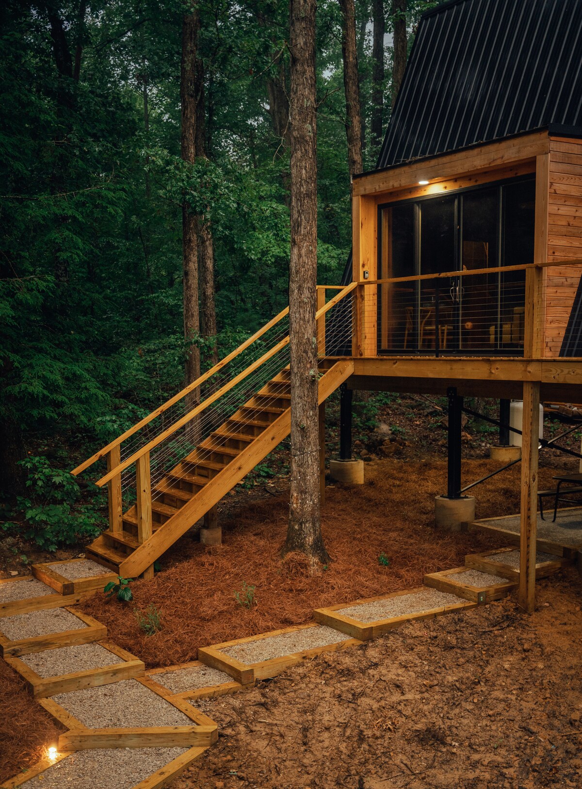 A wooden walkway leads to a set of stairs, guiding visitors to the elevated A-frame cabin. The structure is surrounded by lush trees, and soft lighting illuminates the entrance. Natural materials blend seamlessly with the forest environment, creating a serene and welcoming approach.