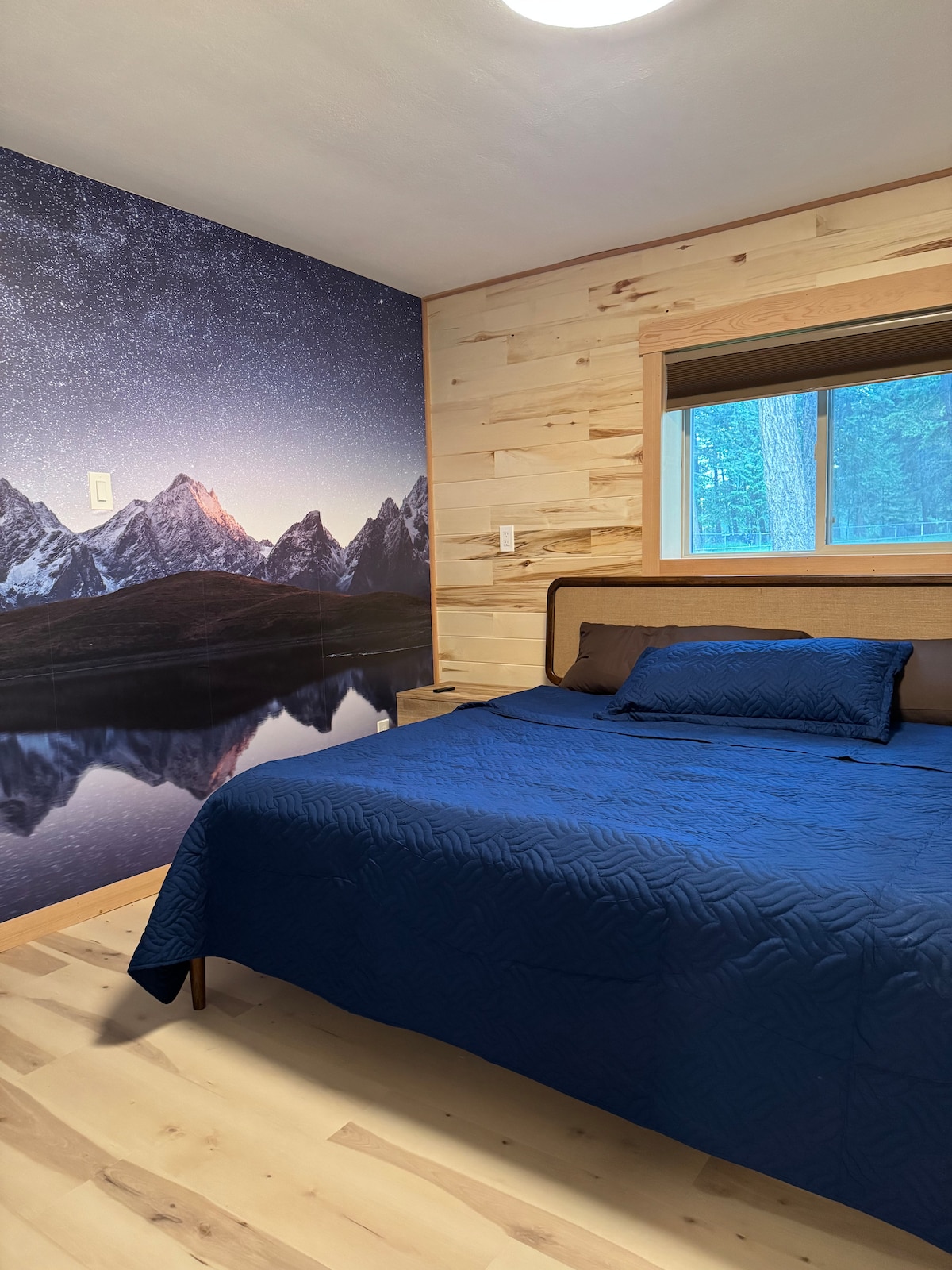 A spacious bed covered in a navy blue quilt is positioned near a window, allowing natural light to enter. A photographic mural of mountain peaks and a reflective lake creates a serene backdrop on one wall. Wooden paneling complements the room's natural aesthetic.